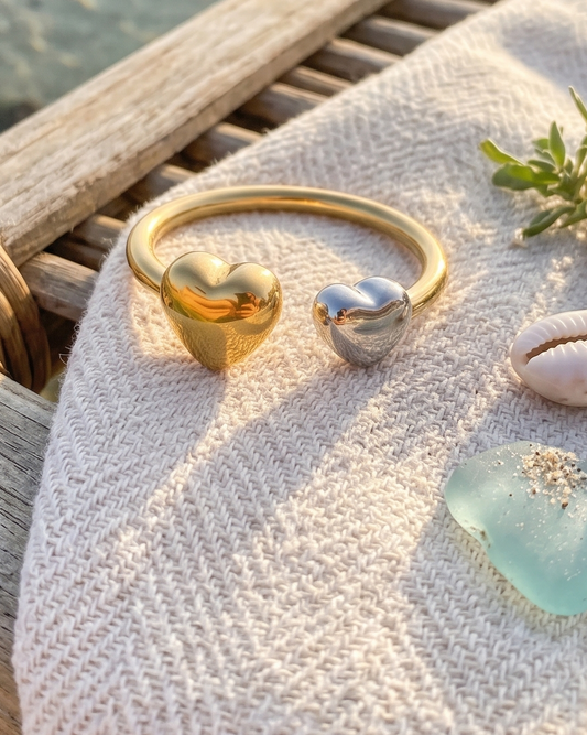 Two heart-shaped cuff rings, one gold and one silver, displayed on a woven beach chair with a textured cream blanket, seashells, sea glass, and small succulents, with a tide pool and sandy beach in the background.