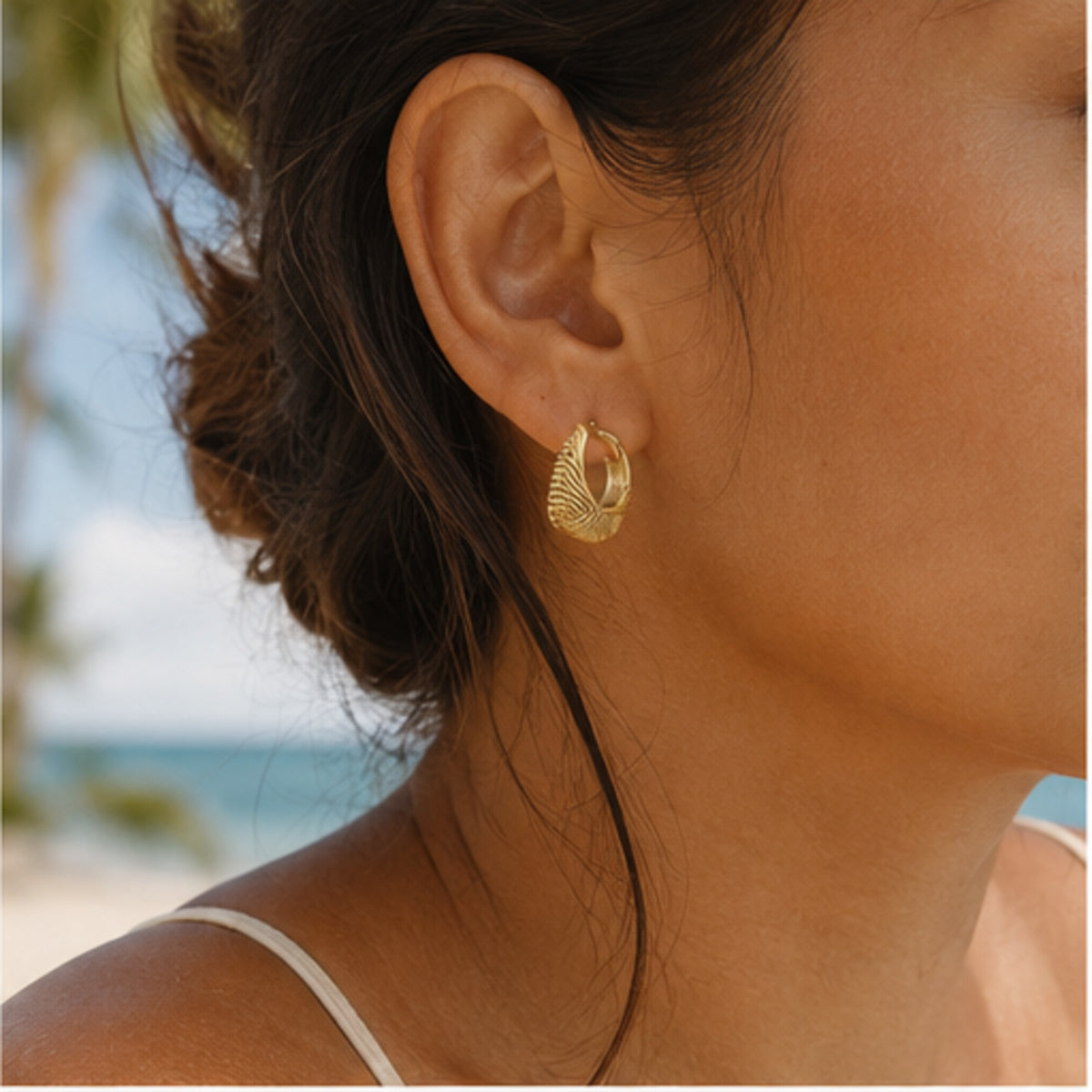 A close-up of a woman's ear wearing a textured gold hoop earring, with a blurred beach and ocean background.
