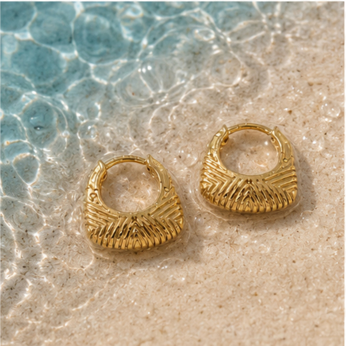 A pair of textured gold huggie hoop earrings resting on wet sand near shallow water, with ripples and blue-green water visible around them.