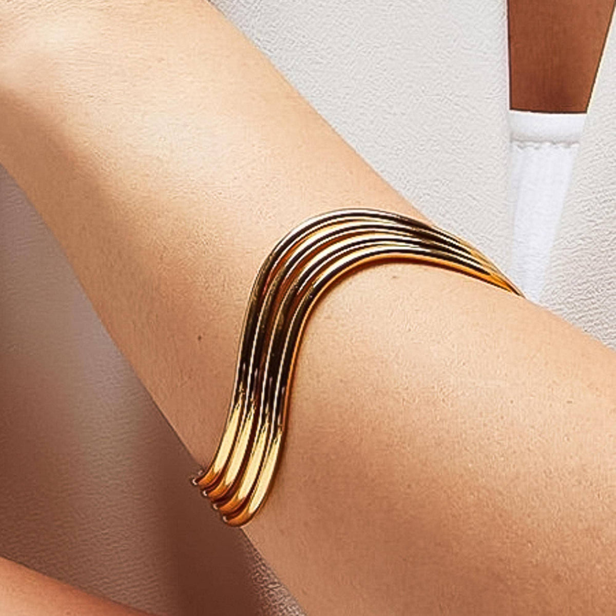 A close-up of a person's forearm wearing a multi-strand metallic cuff bracelet with a wavy, layered design in a gold finish.