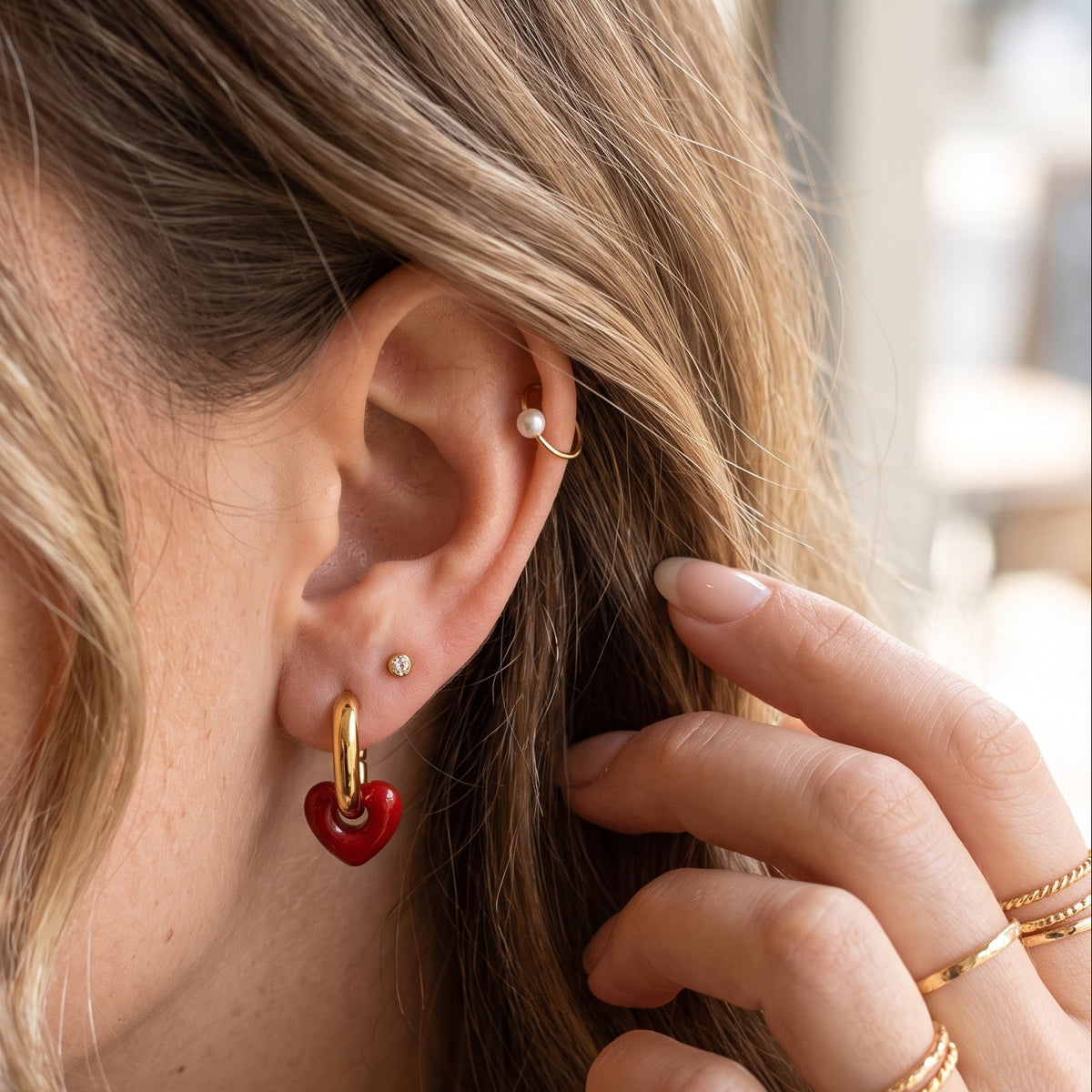 A close-up lifestyle shot of a woman wearing a small gold hoop earring with a dangling red heart charm. She is shown in profile with her hand near her face, wearing other gold rings and ear piercings in a softly lit indoor setting.
