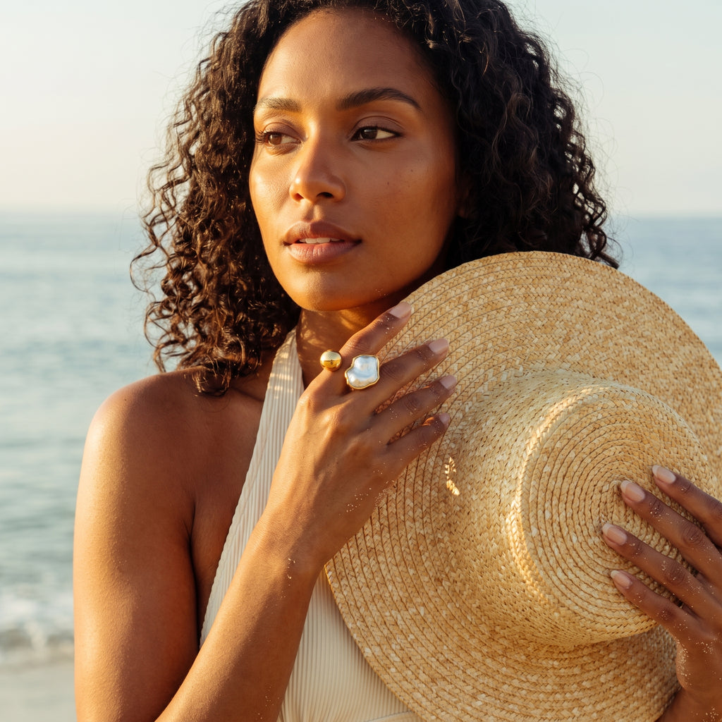 A woman on a beach holds a large woven straw hat while wearing a white halter top and a statement ring with a large white stone.