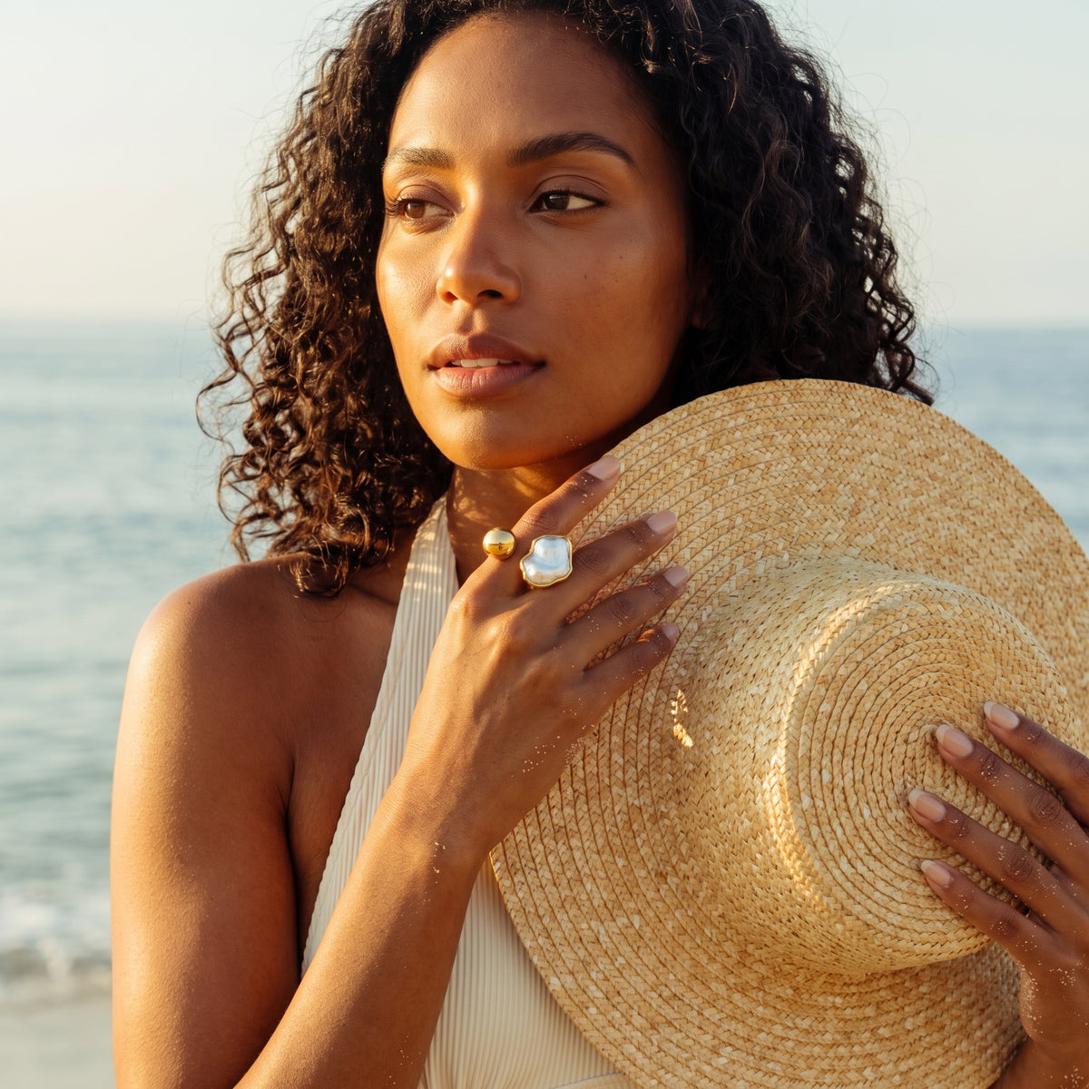 A woman on a beach holds a large woven straw hat while wearing a white halter top and a statement ring with a large white stone.