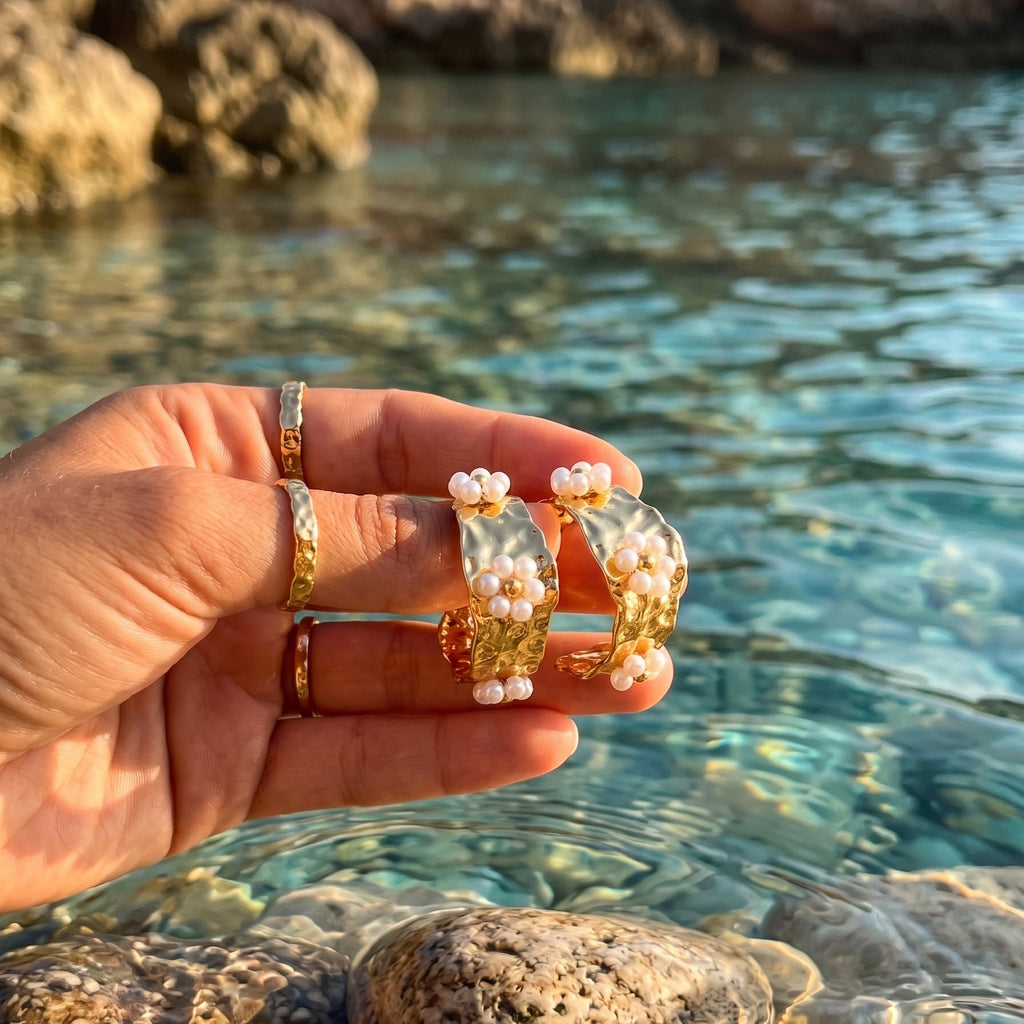 A hand holds a pair of gold hoop earrings decorated with white faux pearls, set against a natural background of clear blue water and rocks.