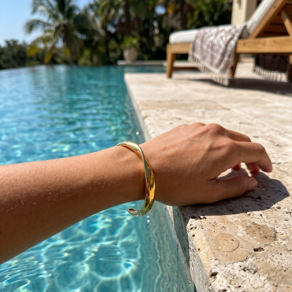 A gold-toned twisted bangle bracelet worn on a person's wrist, shown by the poolside with a stone deck, palm trees, and a lounge chair in the background.