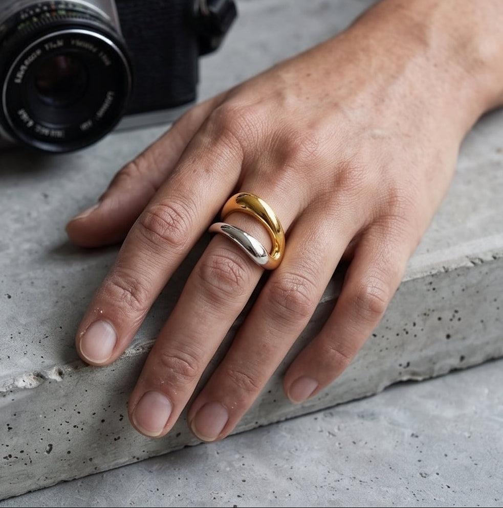 A four-panel collage showing a pair of gold rings worn on hands in different settings: on a wooden desk with a mug, holding a martini glass, resting on a concrete surface with a camera, and on the chest over a sweater.