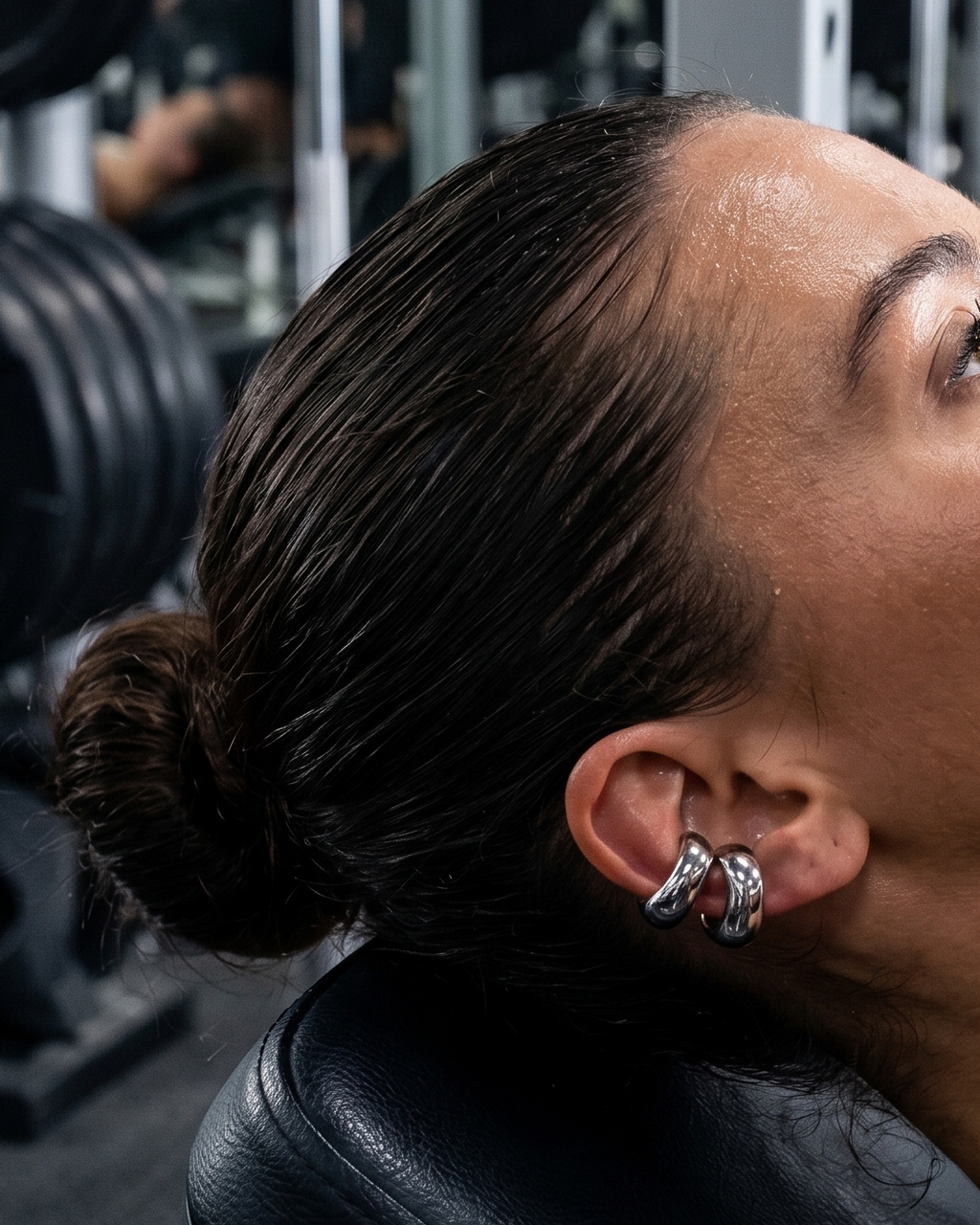 A woman in a gym setting, lying on a weight bench and holding a barbell, wearing silver hoop earrings and a delicate necklace.