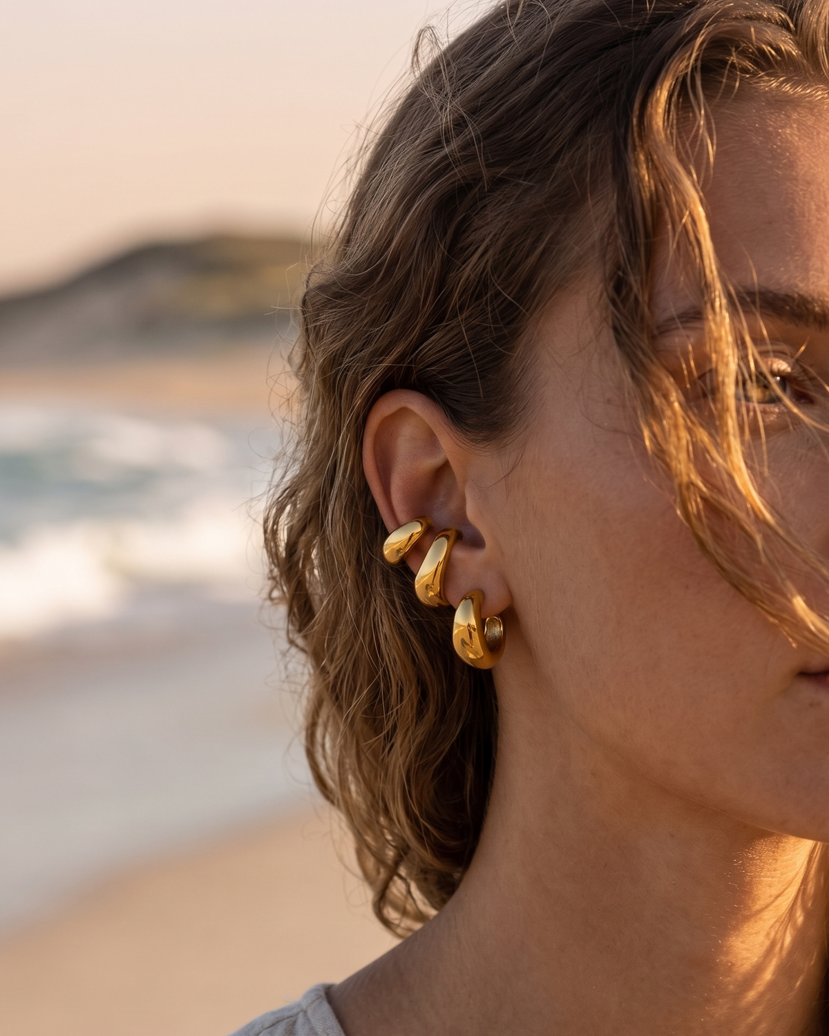 A close-up of a woman on a beach wearing a pair of gold hoop earring cuffs, warm sunset light on her face and hair.