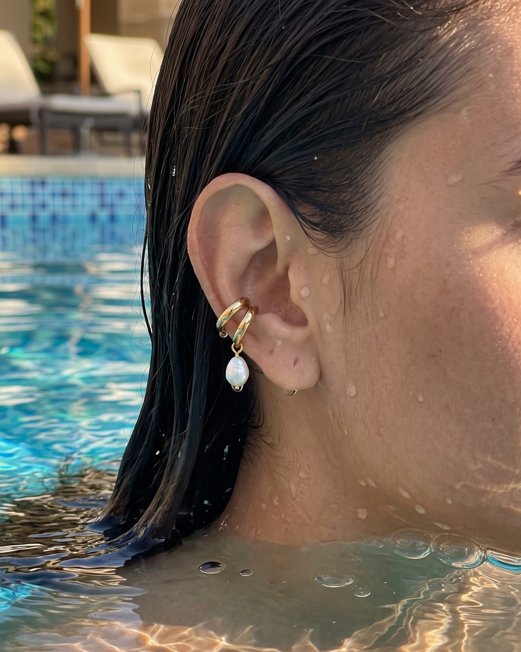 A close-up of a woman's ear wearing a gold ear cuff with a dangling white pearl, set against a swimming pool background with water droplets on the skin.