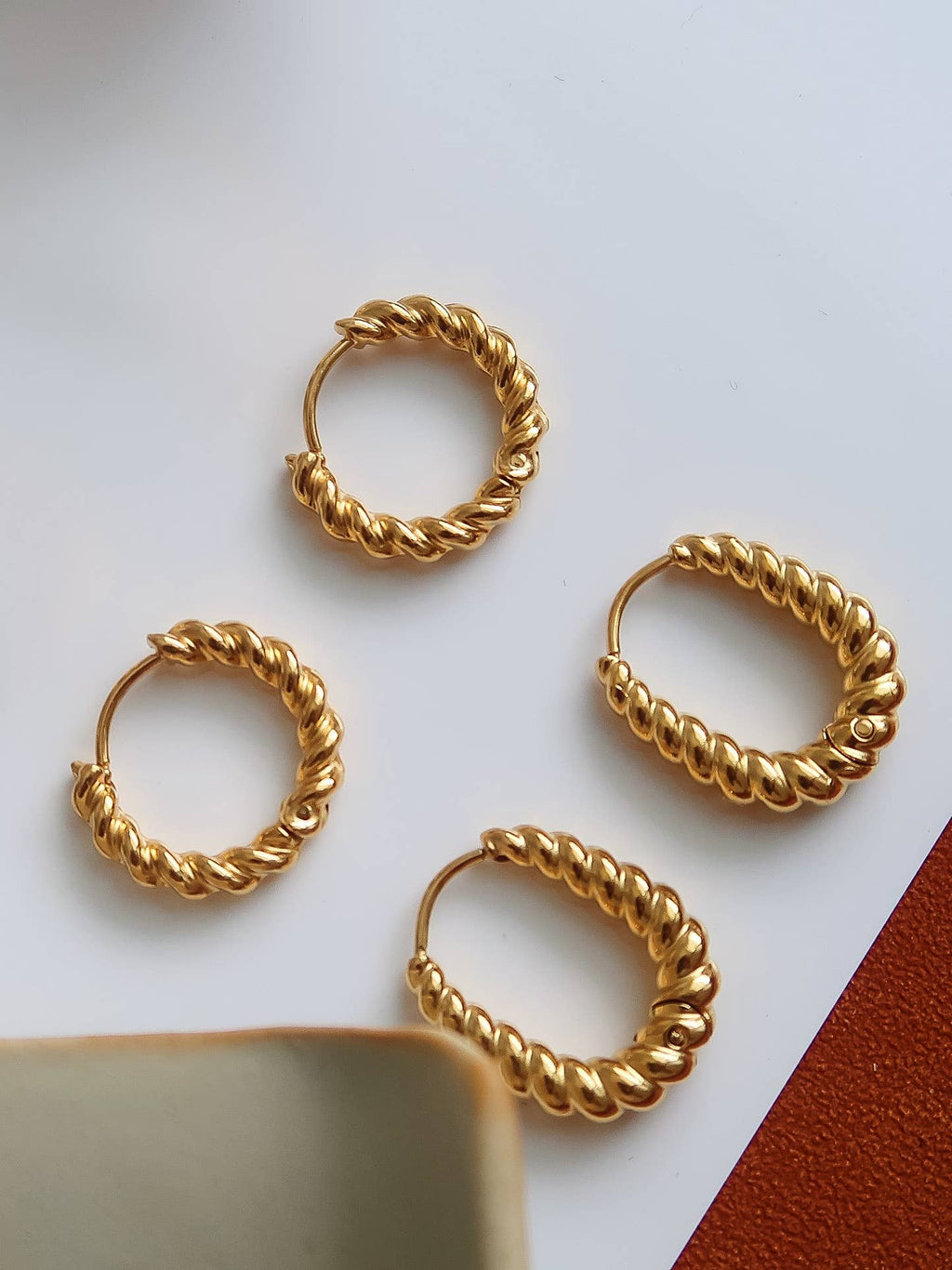 Four twisted gold hoop earrings arranged on a white surface with a small brown textured corner visible in the lower right.
