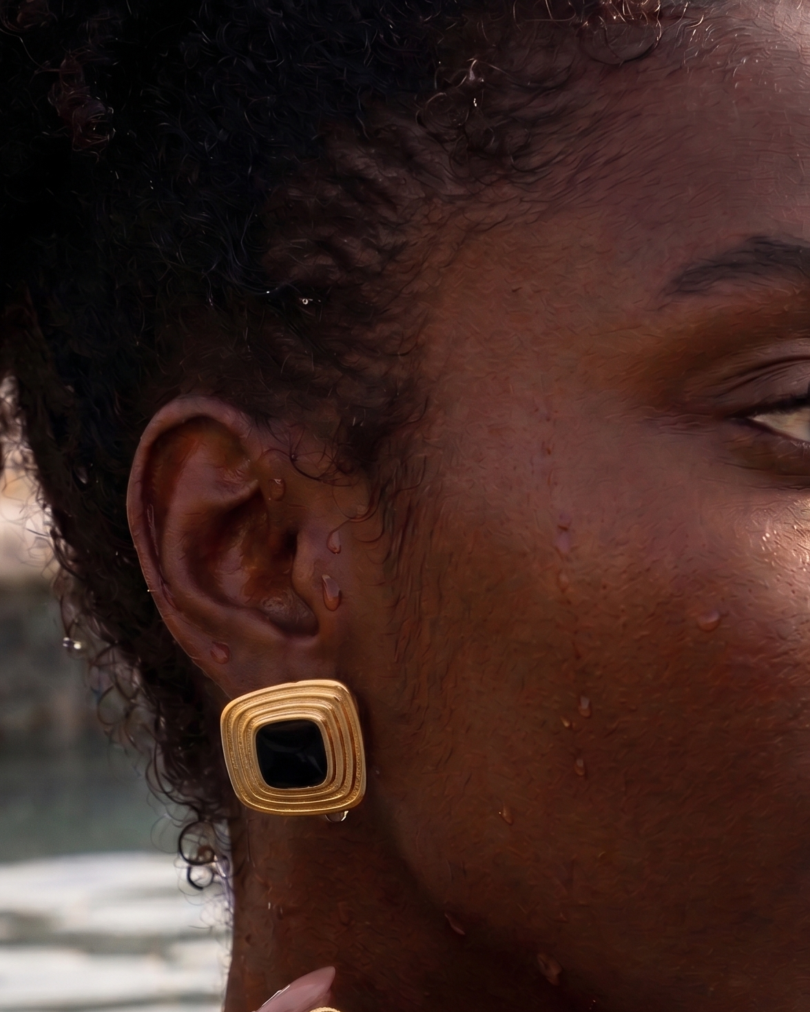 A close-up lifestyle shot of a woman wearing gold square stud earrings with black center stones, set against a warm, sunlit outdoor background near water.
