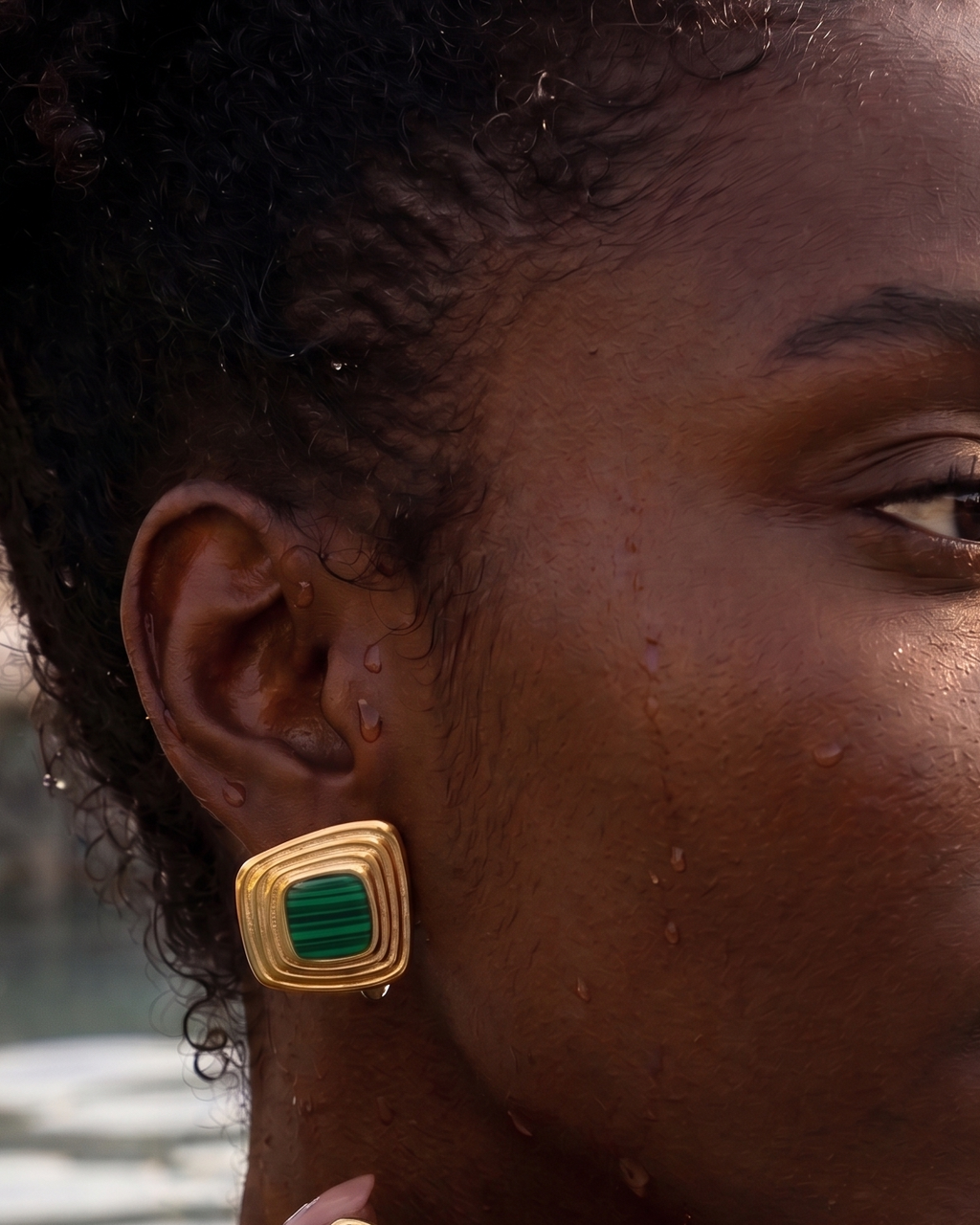 A close-up lifestyle shot of a woman wearing gold square earrings with green inlay, set against a warm, sunlit outdoor background with water.