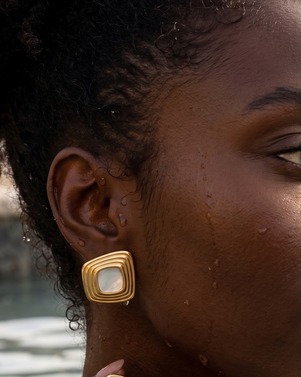 A close-up lifestyle shot of a woman wearing gold square earrings with white inlay, set against a warm, sunlit outdoor background near water.