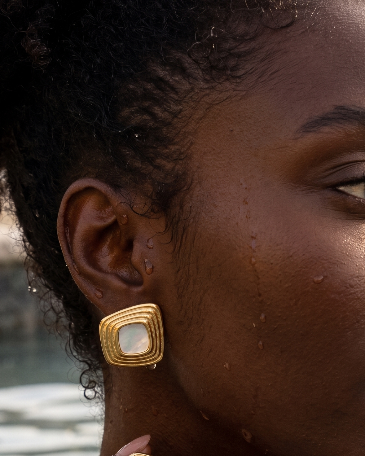 A close-up lifestyle shot of a woman wearing gold square earrings with white inlay, set against a warm, sunlit outdoor background near water.