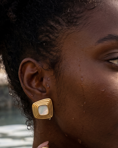 A close-up lifestyle shot of a woman wearing gold square earrings with white inlay, set against a warm, sunlit outdoor background near water.