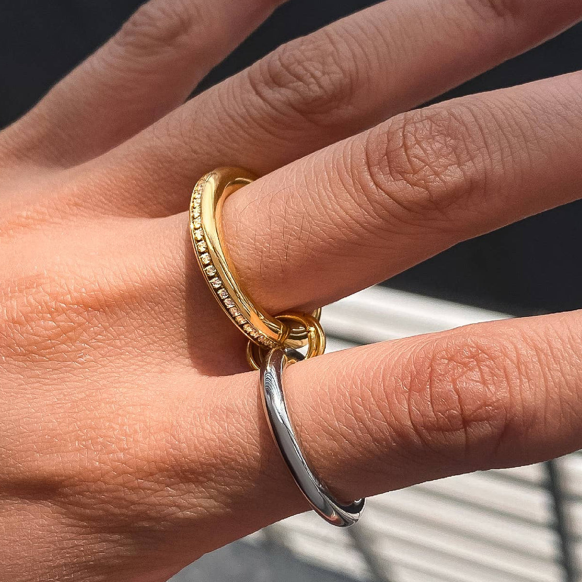 Hand wearing a gold ring with a silver band on a blurred outdoor background
