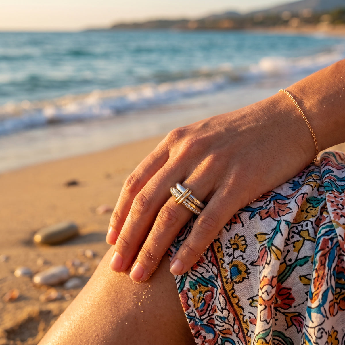 A close-up lifestyle shot of a person's hand resting on their knee at the beach, wearing a double-band ring with a gold-toned center band and a silver-toned outer band with a small white inlay. A delicate gold chain bracelet is also visible on the wrist, with the blue ocean and sandy shore blurred in the background.
