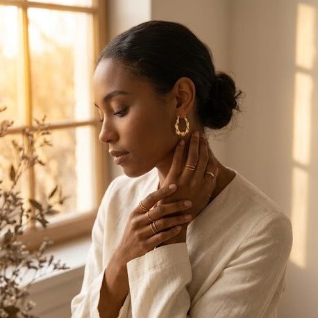 A split lifestyle and product shot featuring gold jewelry, including a gold ring and hoop earrings, displayed on a marble tray next to a small amber dropper bottle. On the right, a woman in a white top is shown wearing the gold hoop earring and touching her neck in a warm, sunlit room.