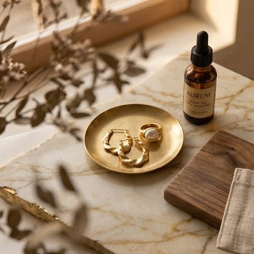 A split lifestyle image showing gold jewelry, including a gold hoop earring worn by a woman on the right and gold rings on a tray with a gold dropper bottle on the left, both styled on a marble tray with dried botanicals in a warm, sunlit room.